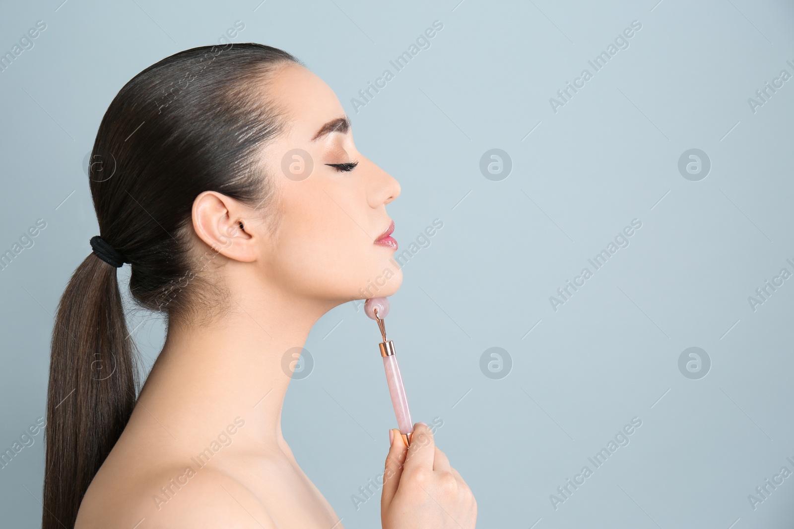 Photo of Woman using natural pink quartz face roller on grey background, space for text
