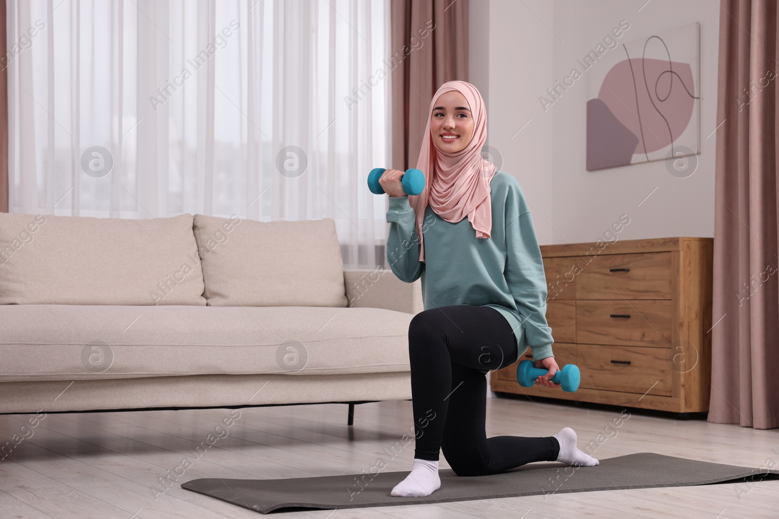 Photo of Muslim woman in hijab doing exercise with dumbbells on fitness mat at home. Space for text