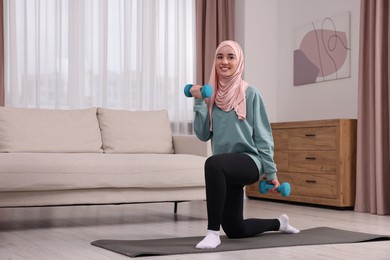 Photo of Muslim woman in hijab doing exercise with dumbbells on fitness mat at home. Space for text
