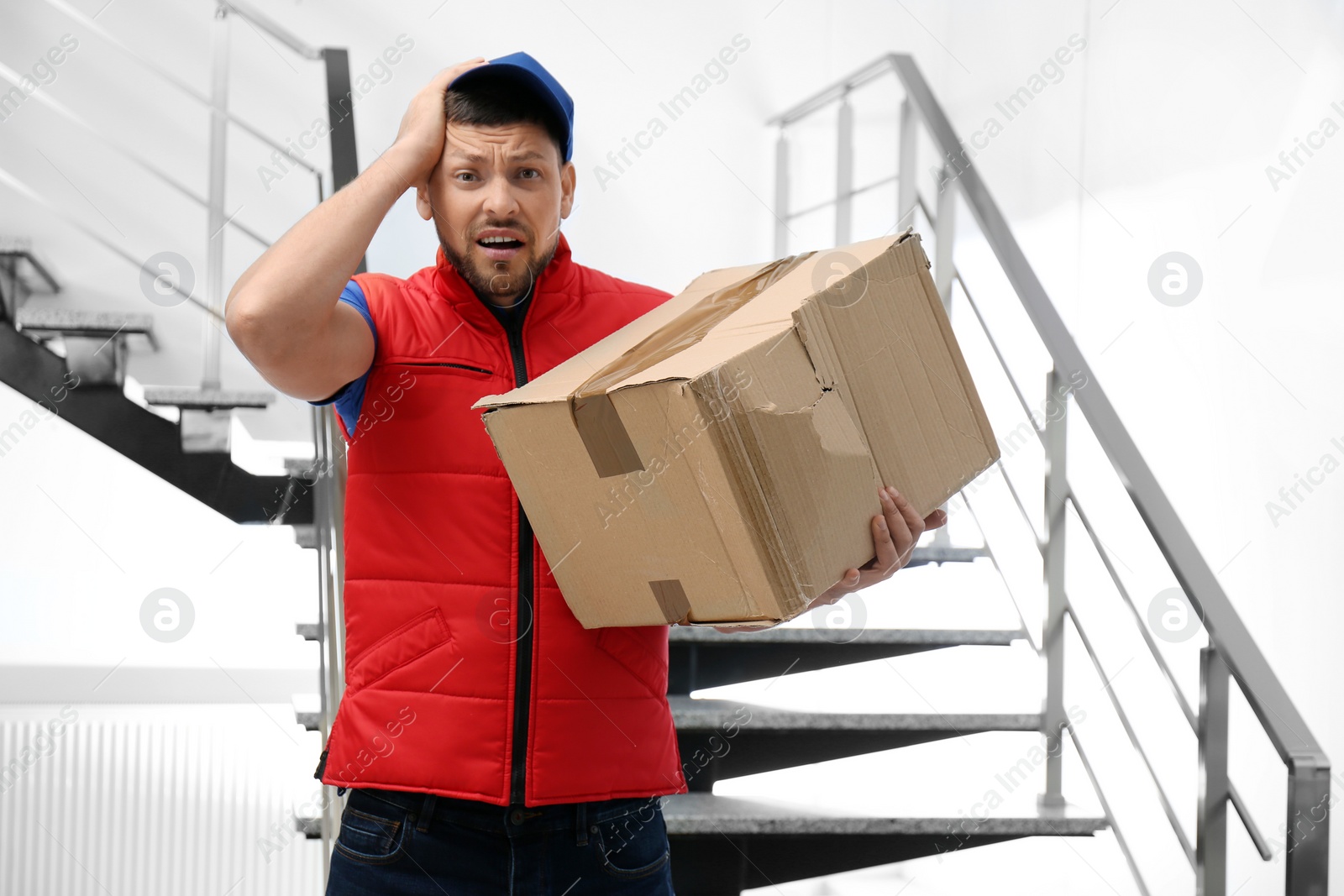 Photo of Emotional courier with damaged cardboard box indoors. Poor quality delivery service