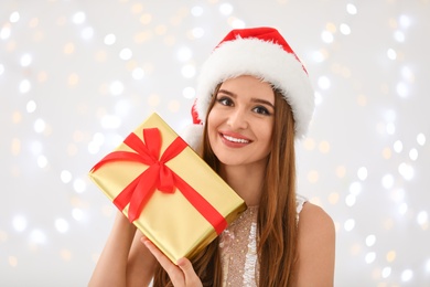 Happy young woman in Santa hat with gift box against blurred Christmas lights