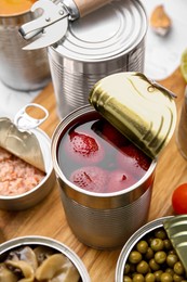 Open tin cans with different preserved products on wooden board, closeup