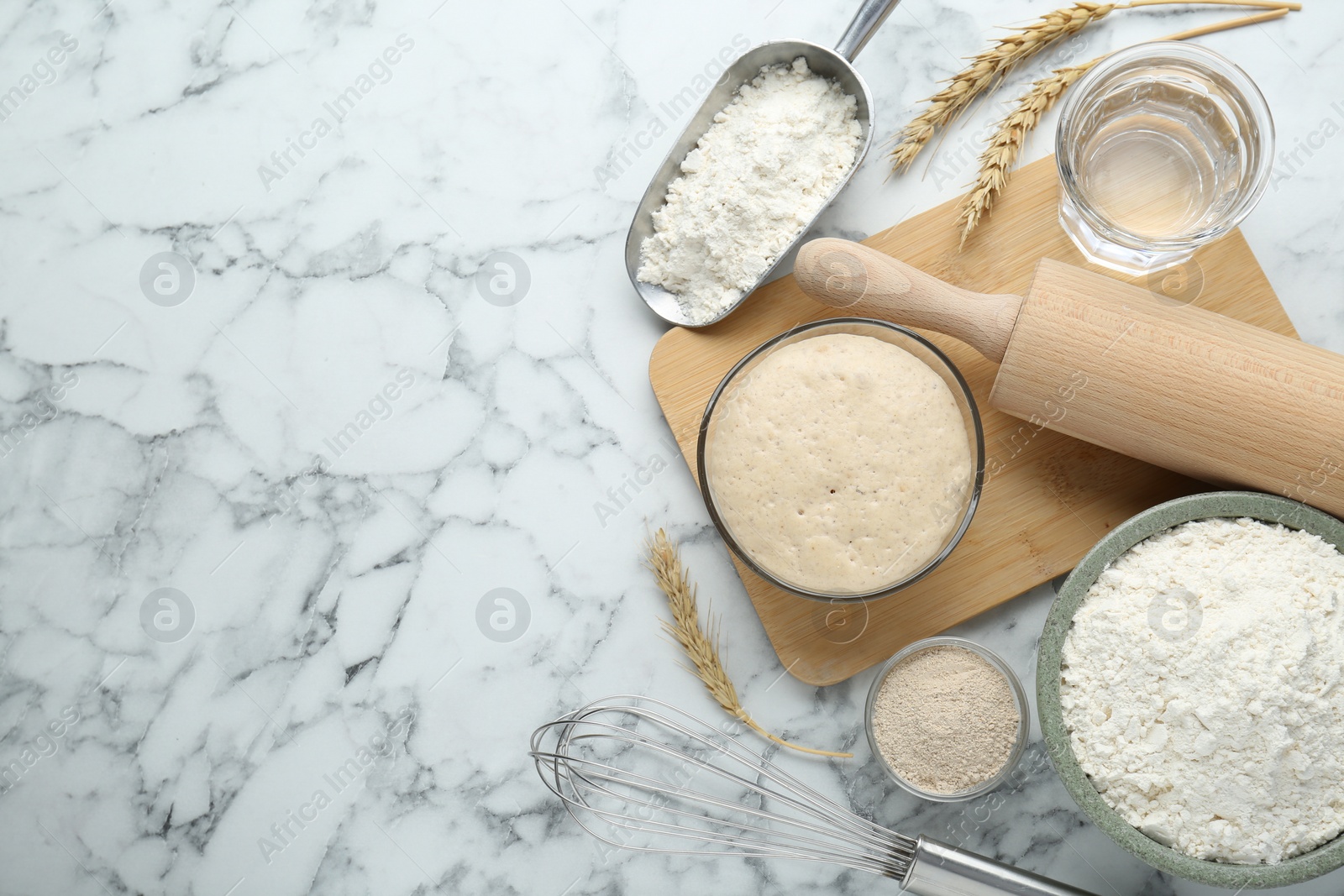 Photo of Leaven, flour, ears of wheat, rolling pin, whisk and water on white marble table, flat lay. Space for text