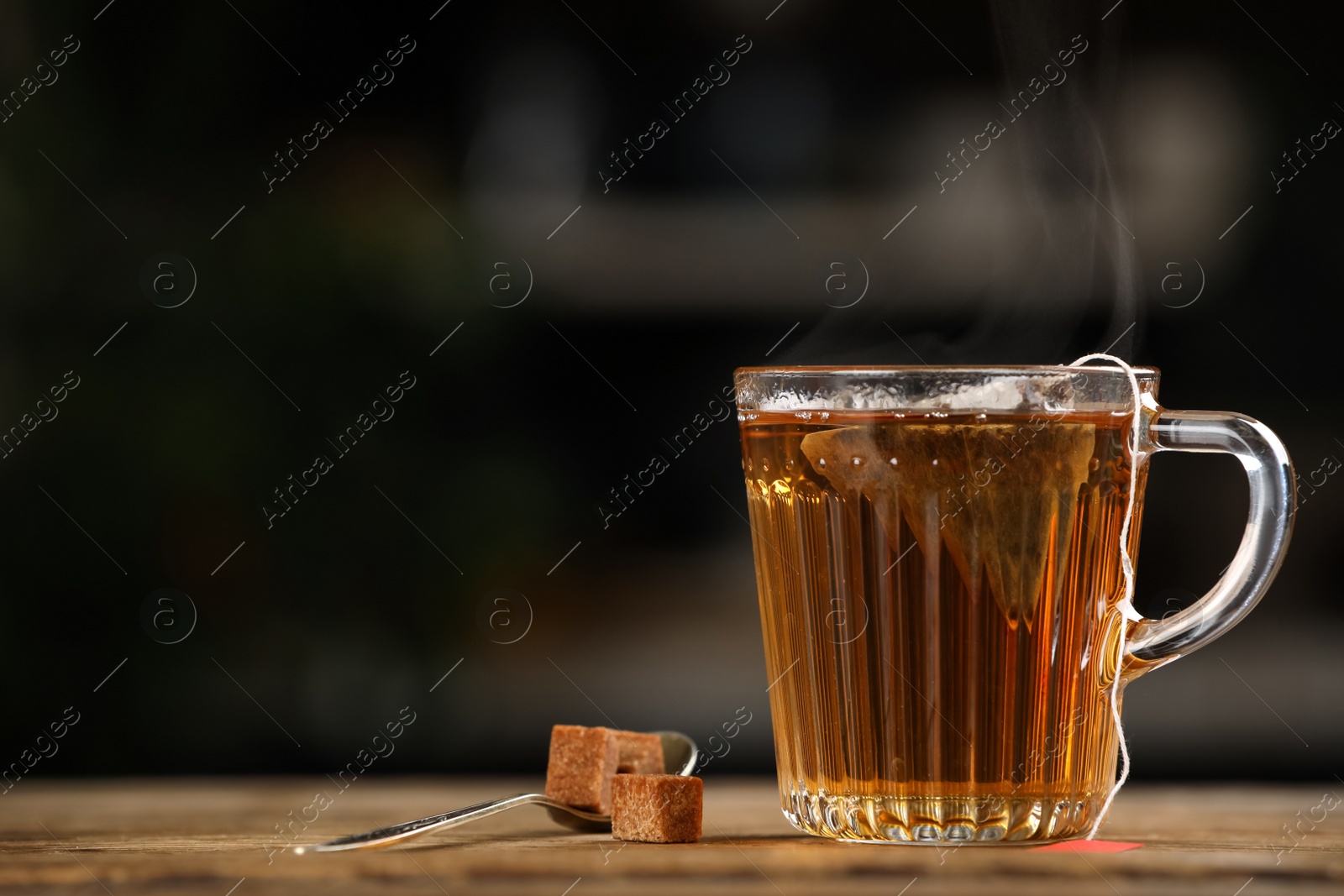 Photo of Tea bag in glass cup of hot water and sugar on wooden table against blurred background