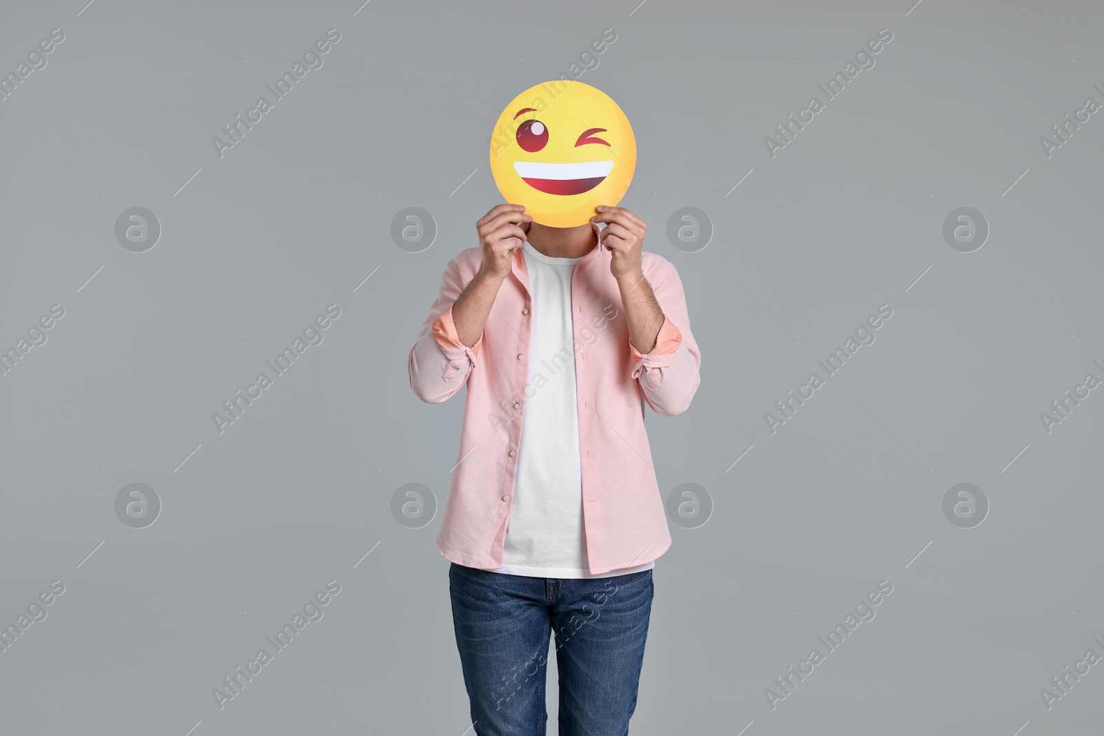 Photo of Man covering face with happy winking emoticon on grey background