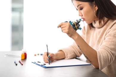 Photo of Professional tattoo artist holding machine and drawing sketch at table indoors