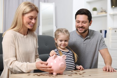 Family budget. Little girl putting coin into piggy bank while her parents watching indoors