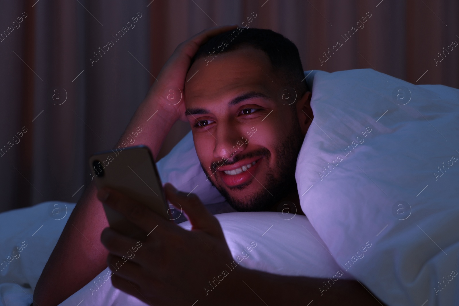 Photo of Happy man using smartphone under blanket in bed at night. Internet addiction