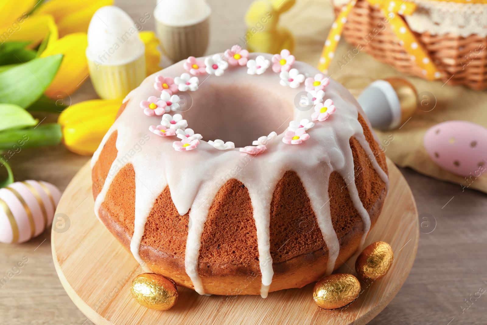 Photo of Delicious Easter cake decorated with sprinkles near painted eggs and tulips on wooden table, closeup