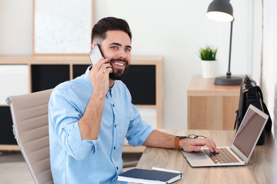 Photo of Handsome young man talking on phone while working at table with laptop in home office