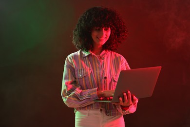 Beautiful young woman with laptop on color background in neon lights and smoke