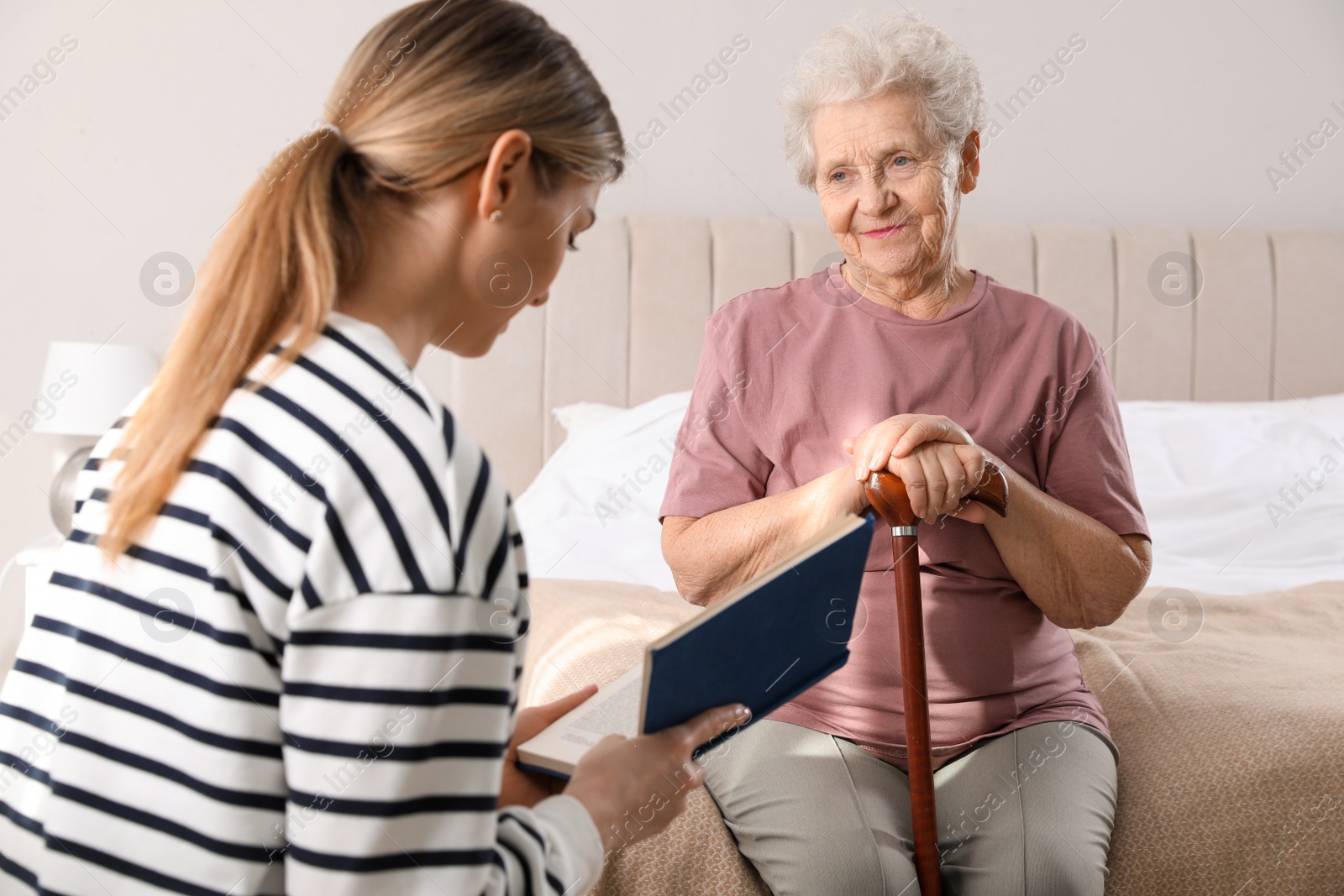 Photo of Young caregiver reading book to senior woman in bedroom. Home care service