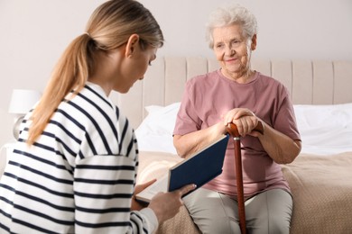 Photo of Young caregiver reading book to senior woman in bedroom. Home care service