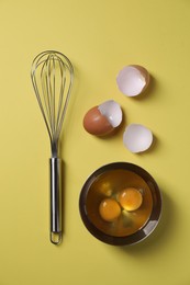 Photo of Metal whisk, raw eggs in bowl and shells on yellow background, flat lay
