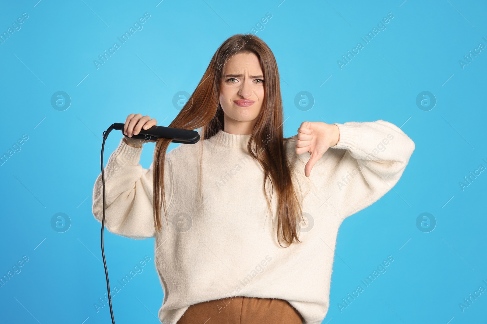 Photo of Upset young woman with flattening iron showing thumb down on light blue background. Hair damage