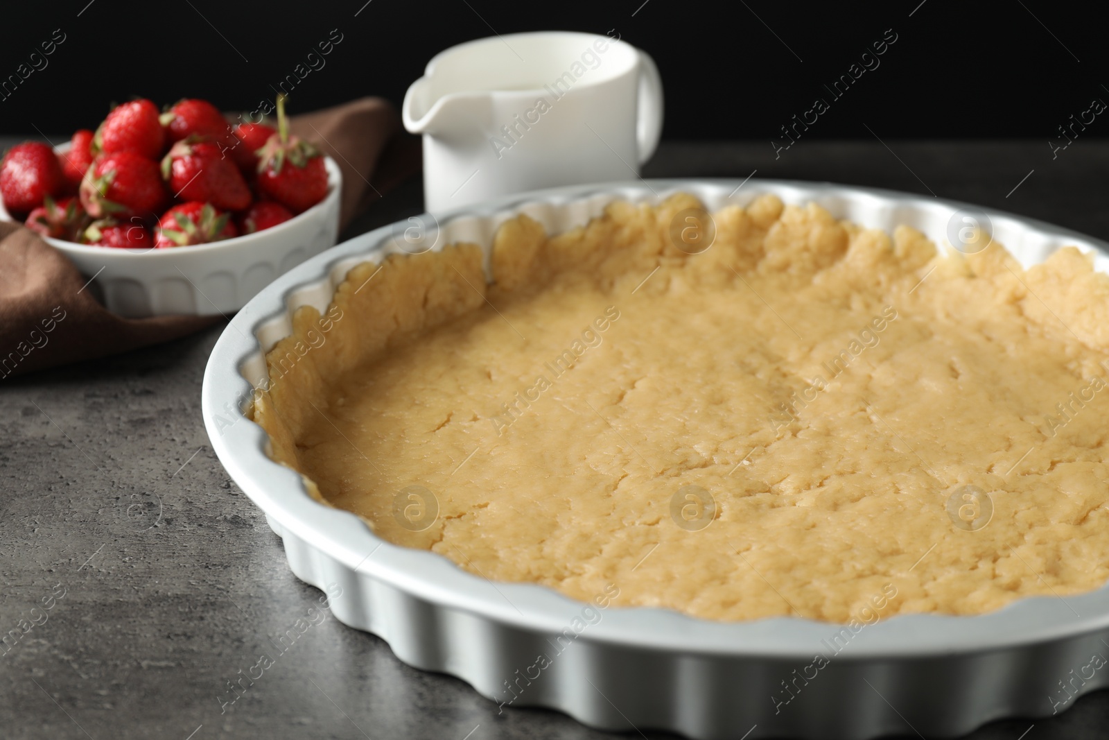 Photo of Making shortcrust pastry. Raw dough in baking dish, strawberries and milk on grey table, closeup