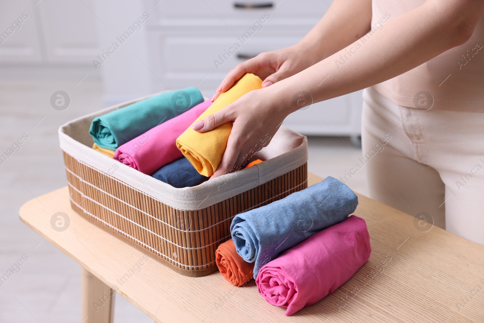 Photo of Woman putting rolled shirt into basket at table in room, closeup. Organizing clothes