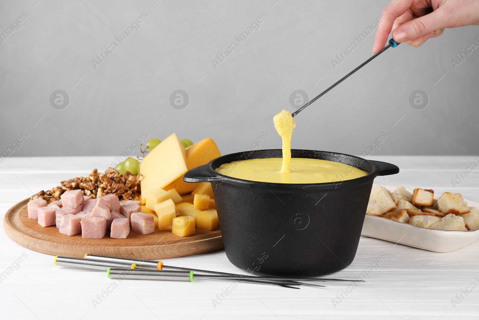 Photo of Woman dipping piece of ham into fondue pot with tasty melted cheese at white wooden table, closeup