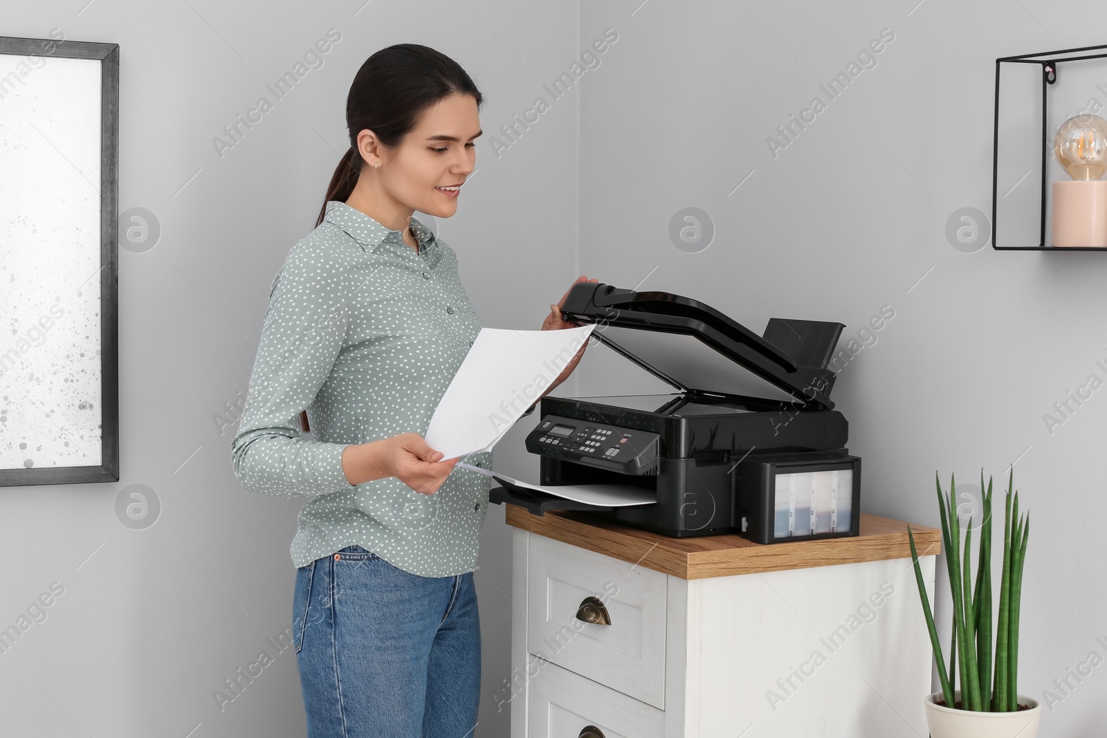 Photo of Woman using modern printer on chest of drawers at home