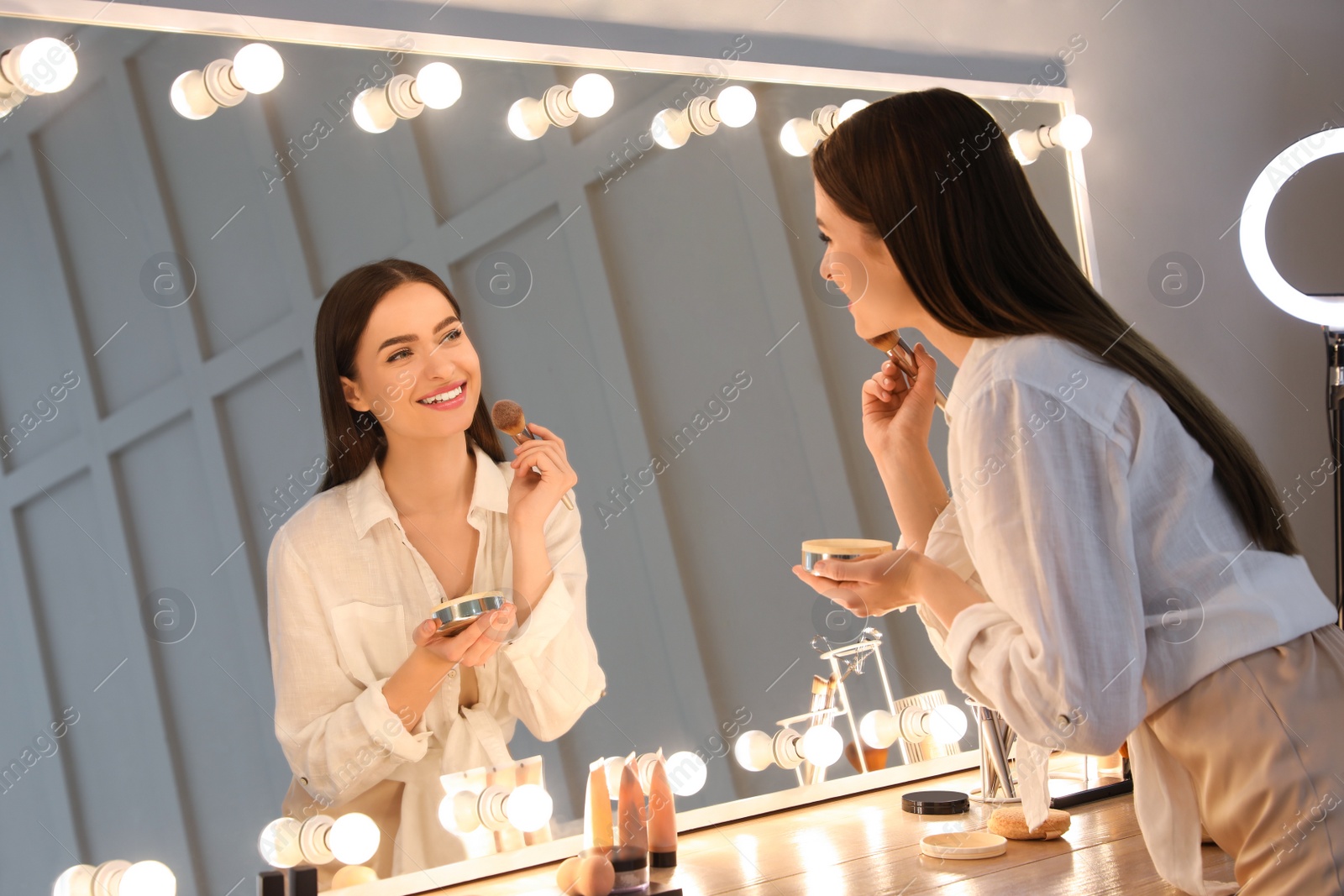 Photo of Young woman applying make up near illuminated mirror indoors