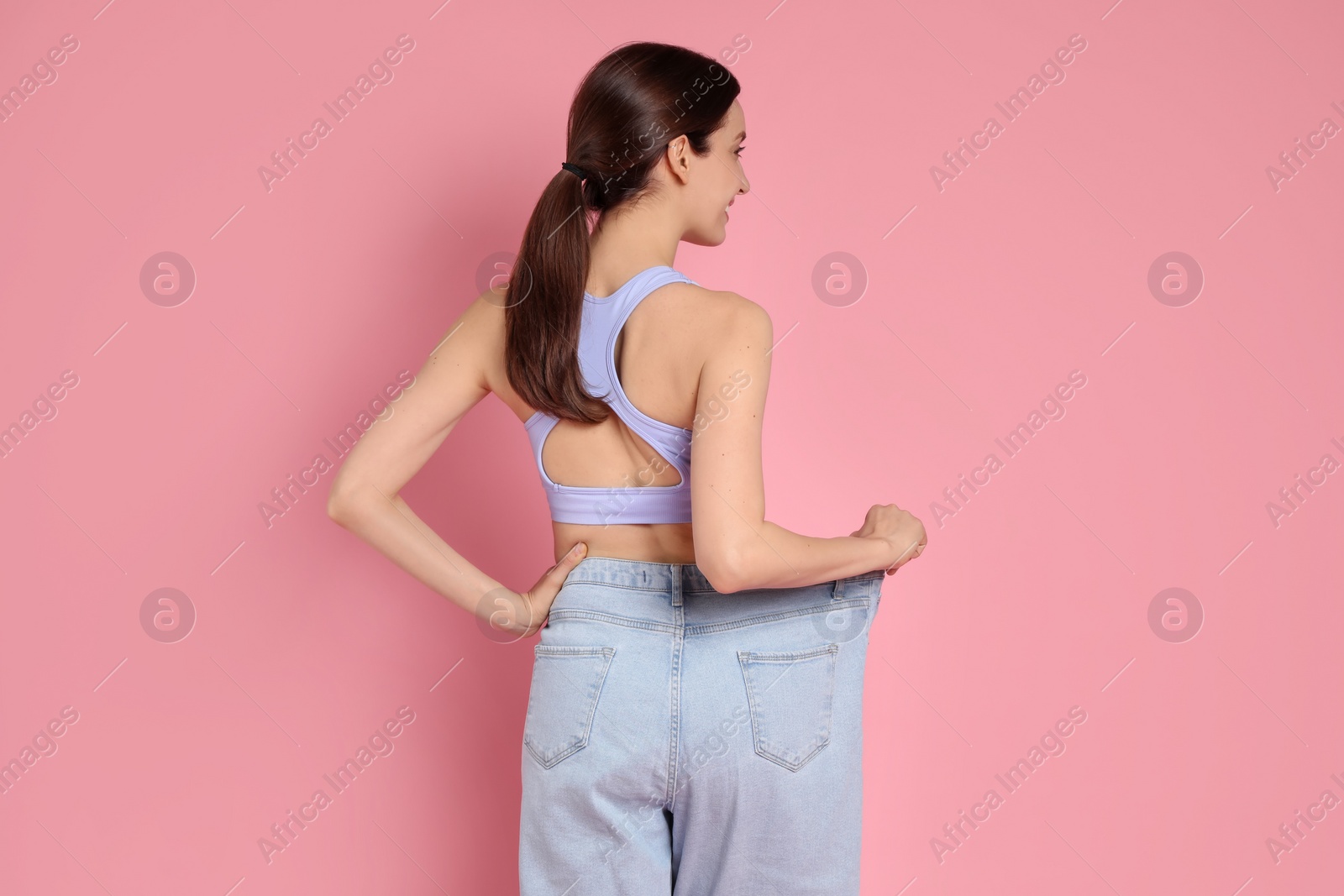 Photo of Woman in big jeans showing her slim body on pink background, back view