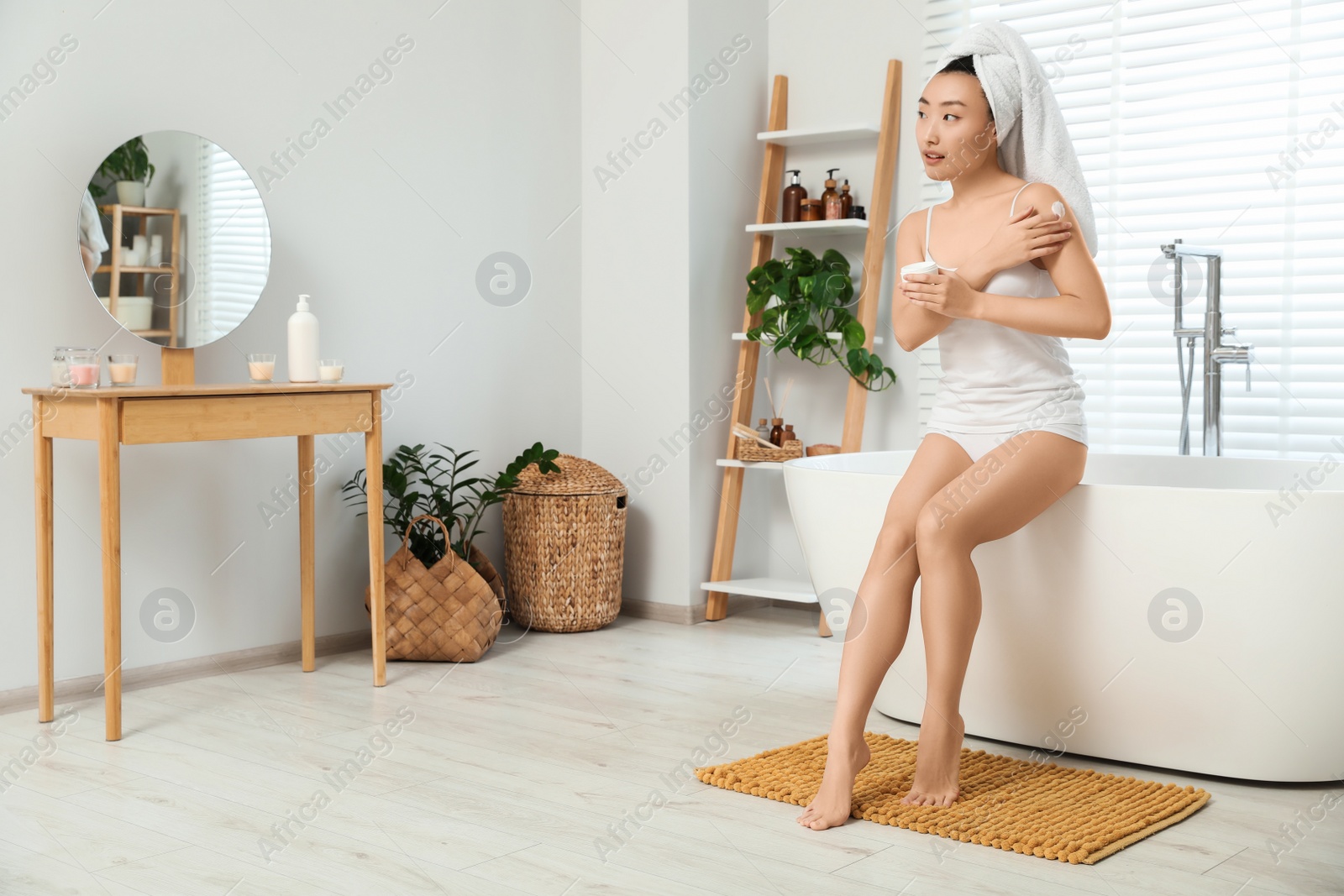 Photo of Beautiful young Asian woman applying body cream on shoulder in bathroom, space for text