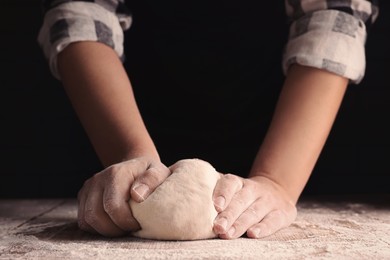 Man kneading dough at wooden table on dark background, closeup