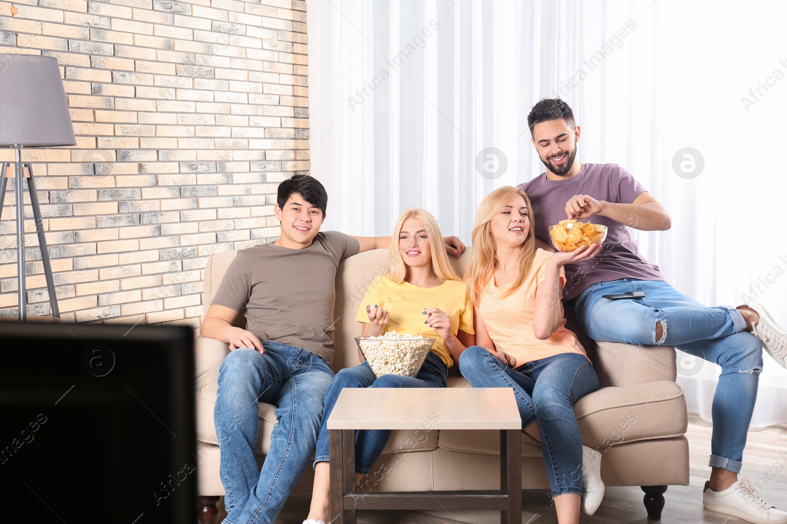 Photo of Young friends with snacks watching TV on sofa at home