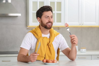 Photo of Happy man holding knife and fork with sausage at table in kitchen