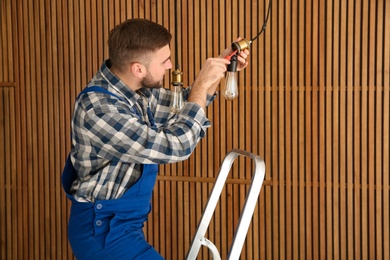 Electrician with screwdriver repairing ceiling lamp against wooden background. Space for text