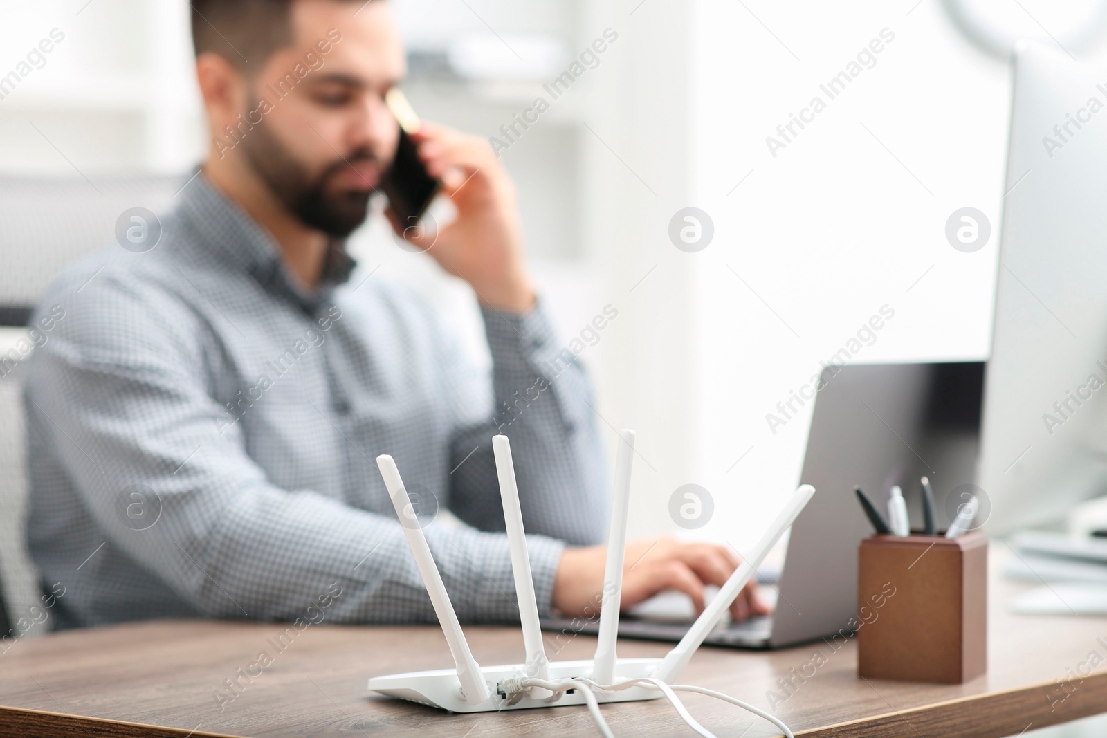 Photo of Man talking on smartphone while working at wooden table indoors, focus on Wi-Fi router