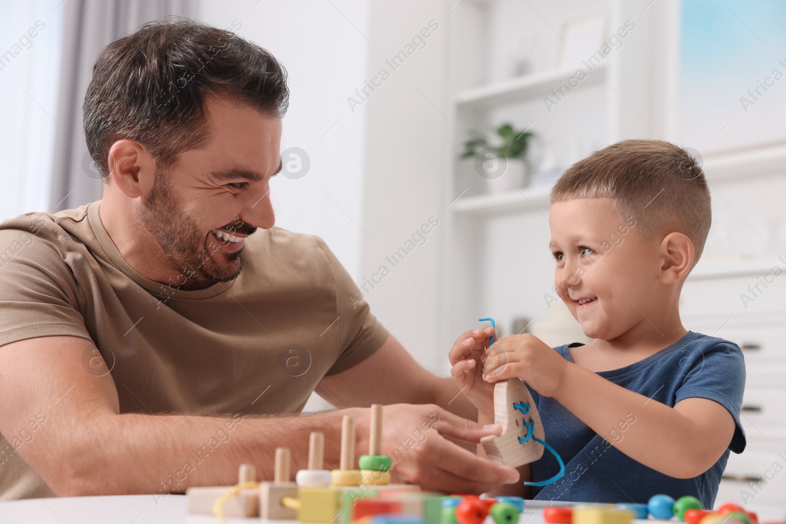 Photo of Motor skills development. Father and his little son playing with wooden lacing toy at white table indoors