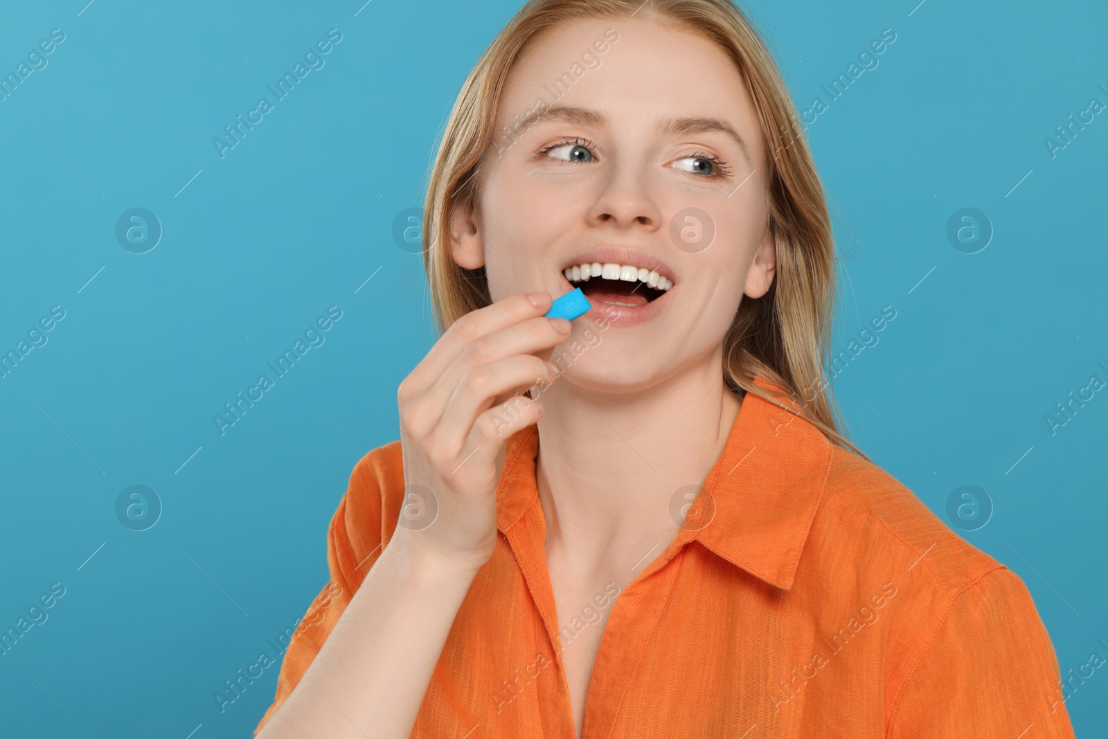 Photo of Happy young woman putting bubble gum into mouth on light blue background