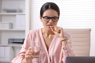 Young woman with pill and glass of water suffering from headache at table in office