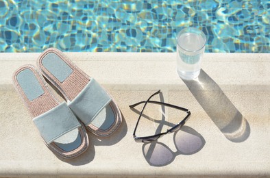 Stylish sunglasses, slippers and glass of water at poolside on sunny day. Beach accessories