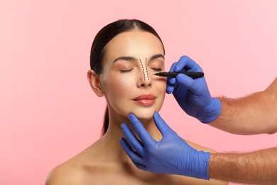 Woman preparing for cosmetic surgery, pink background. Doctor drawing markings on her face, closeup