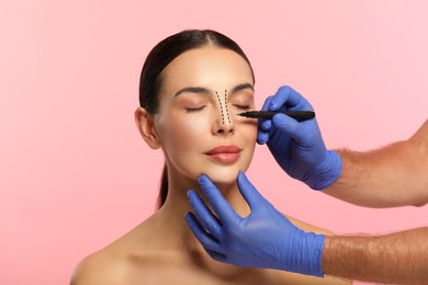 Image of Woman preparing for cosmetic surgery, pink background. Doctor drawing markings on her face, closeup