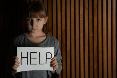 Photo of Sad little girl with sign HELP on wooden background, space for text. Child in danger