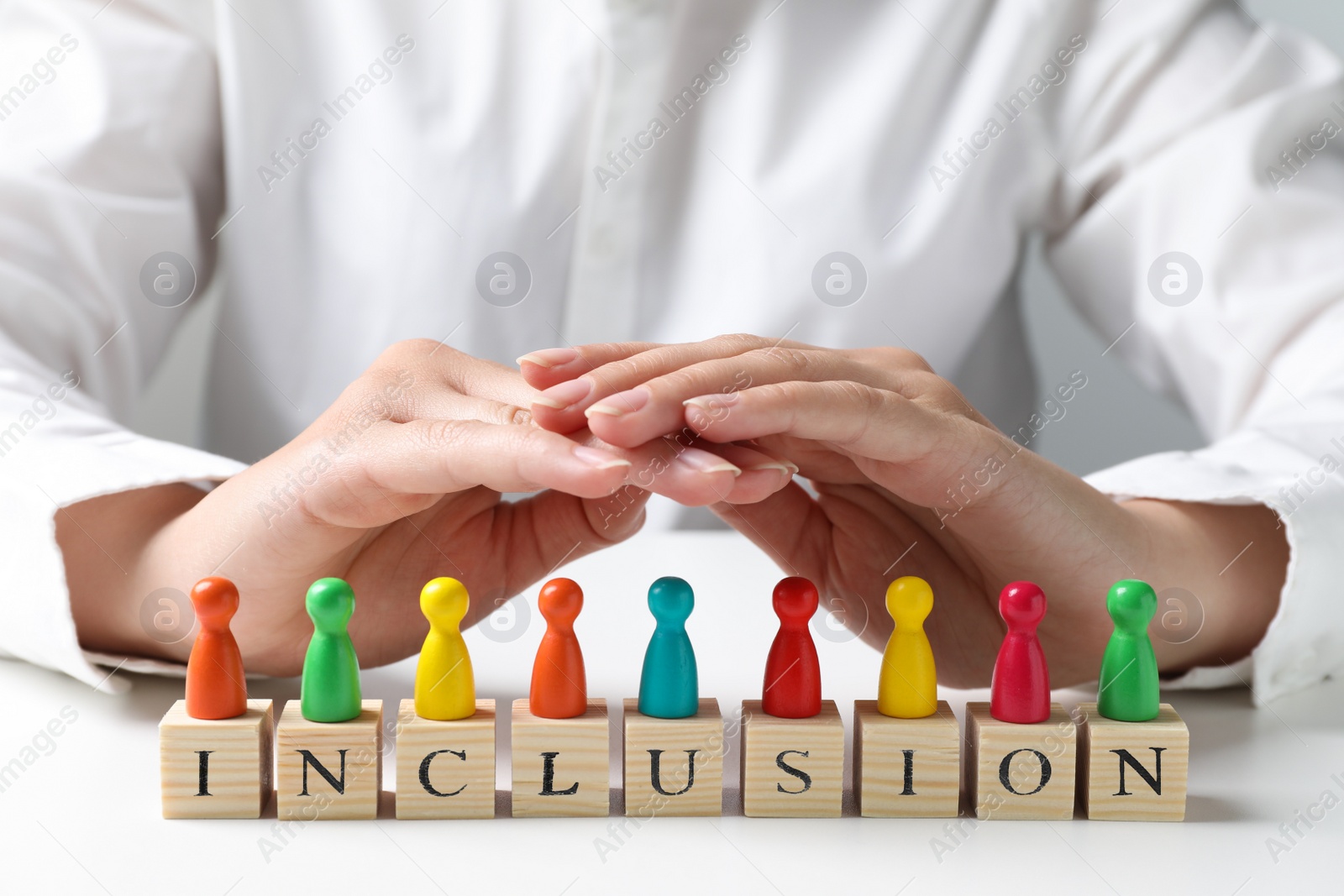 Photo of Woman protecting colorful pawns and wooden cubes with word Inclusion at white table, closeup