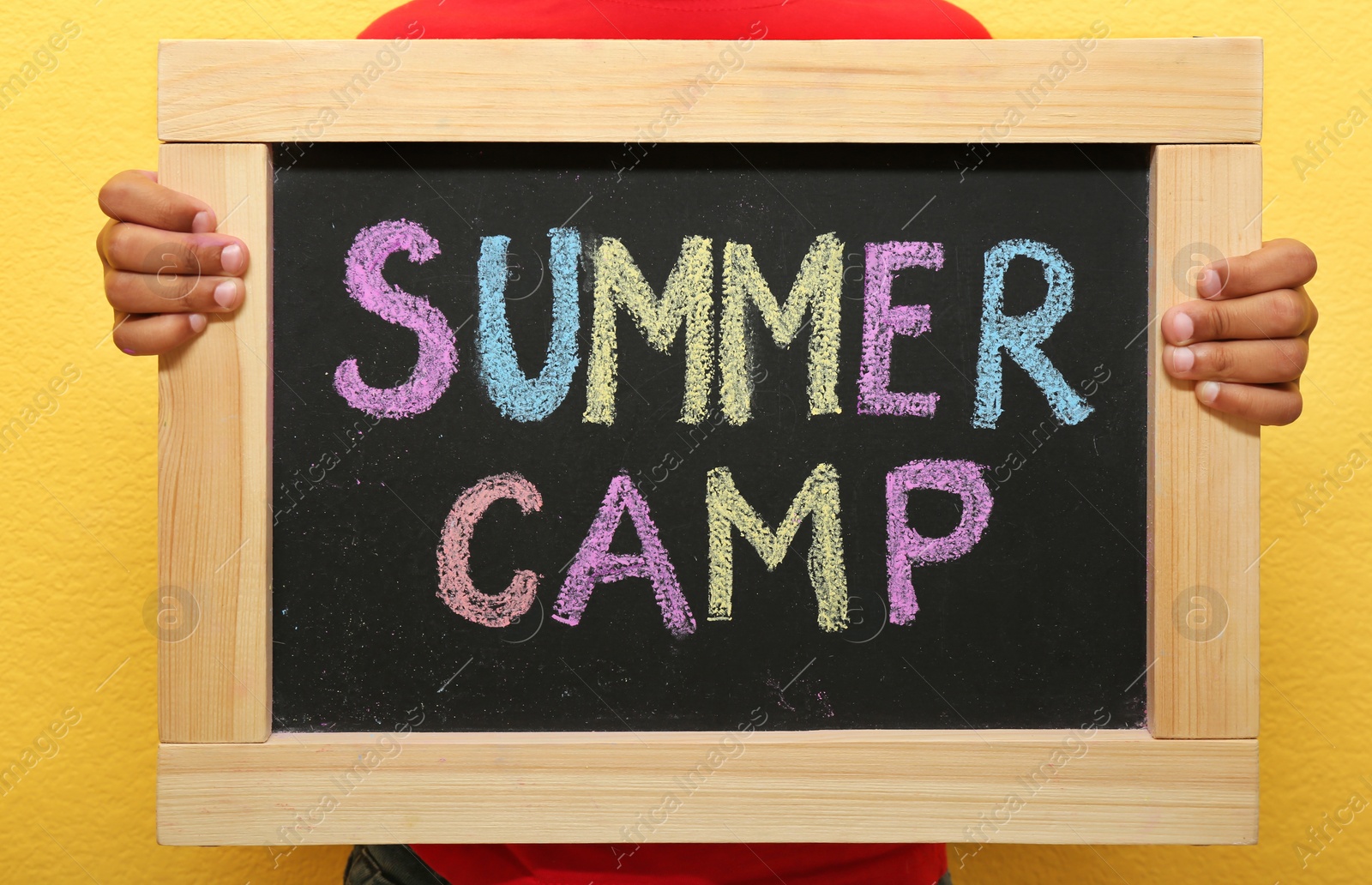 Photo of Child holding blackboard with words SUMMER CAMP chalked in different colors, closeup