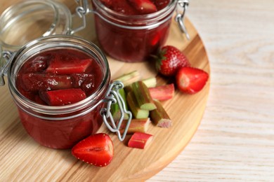 Photo of Jars of tasty rhubarb jam, cut stems and strawberries on white wooden table. Space for text