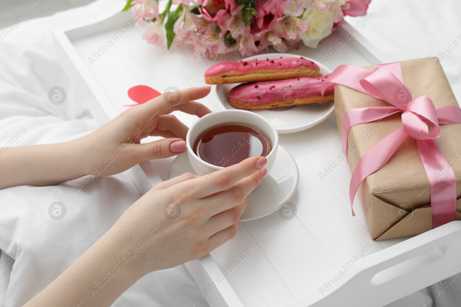 Photo of Tasty breakfast served in bed. Woman with tea, eclairs, gift box and flowers at home, closeup