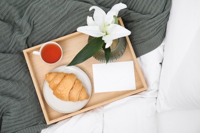 Photo of Tray with tasty croissant, cup of tea and flower on bed, top view