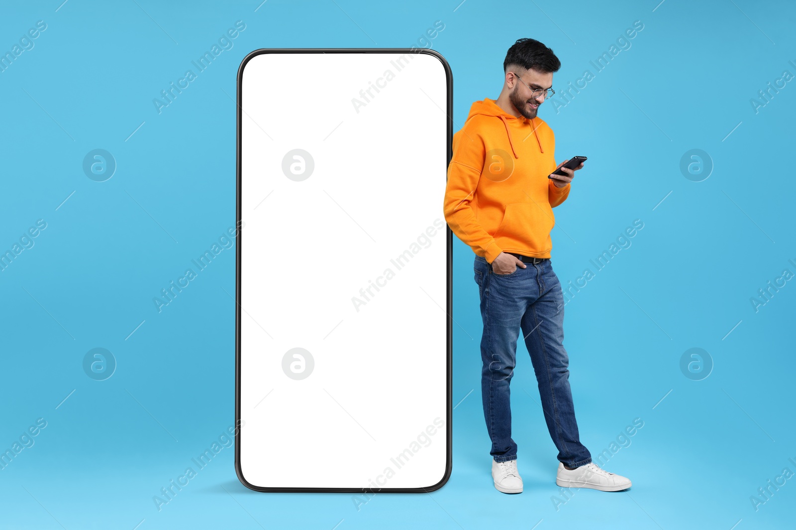 Image of Man with mobile phone standing near huge device with empty screen on light blue background. Mockup for design