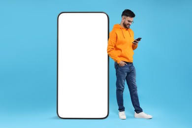 Man with mobile phone standing near huge device with empty screen on light blue background. Mockup for design