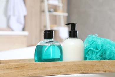 Wooden bath tray with bottles of shower gels and mesh pouf on tub indoors, closeup