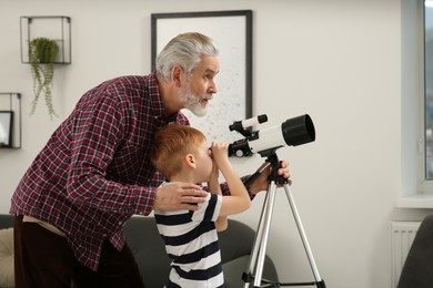 Photo of Little boy with his grandfather looking at stars through telescope in room