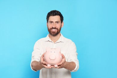 Photo of Happy man with ceramic piggy bank on light blue background