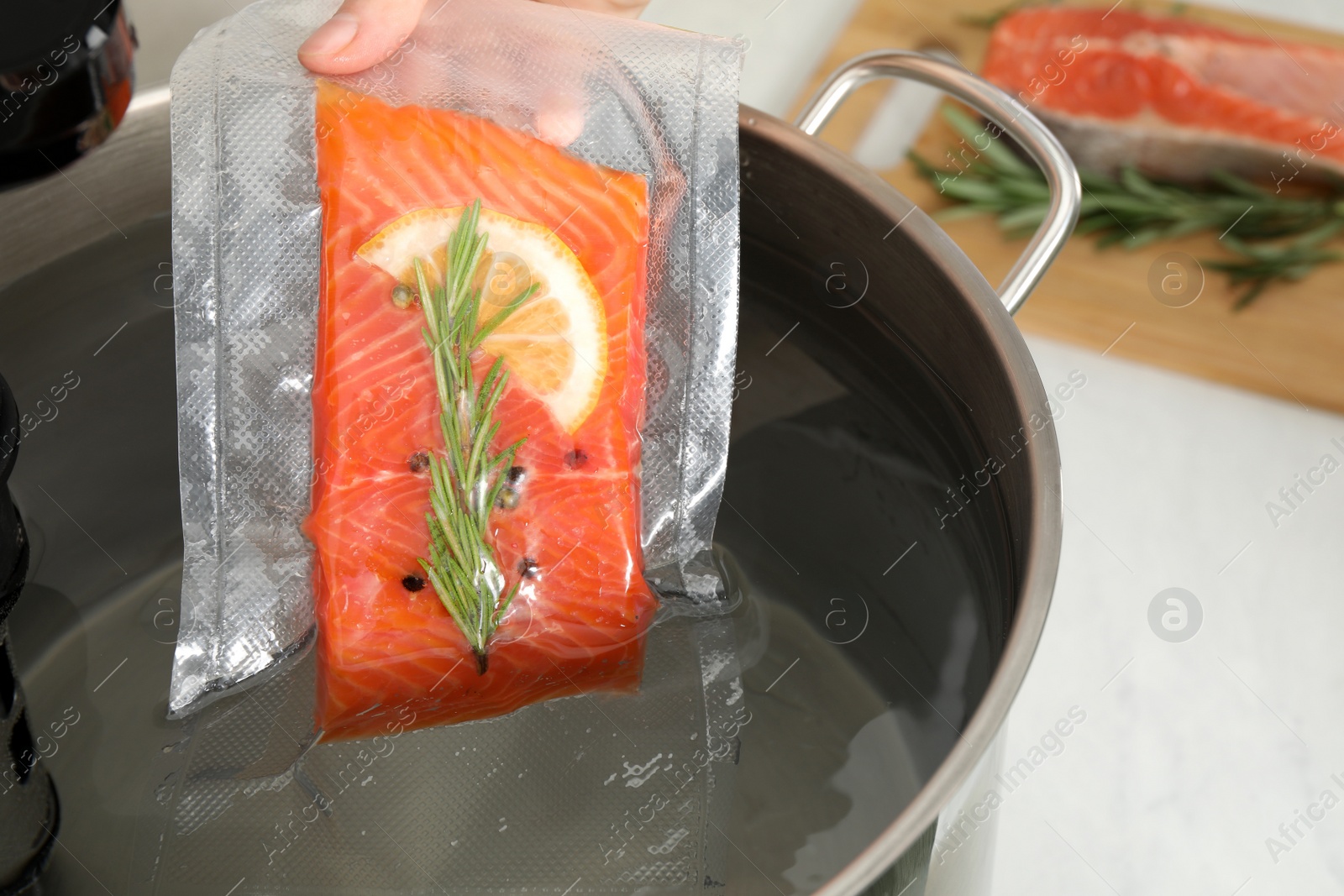 Photo of Woman putting vacuum packed salmon into pot with sous vide cooker in kitchen, closeup. Thermal immersion circulator