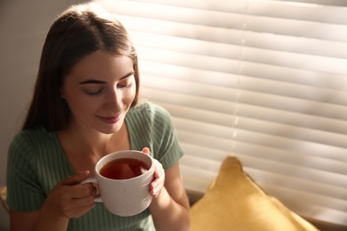 Photo of Beautiful young woman with cup of tea near window at home